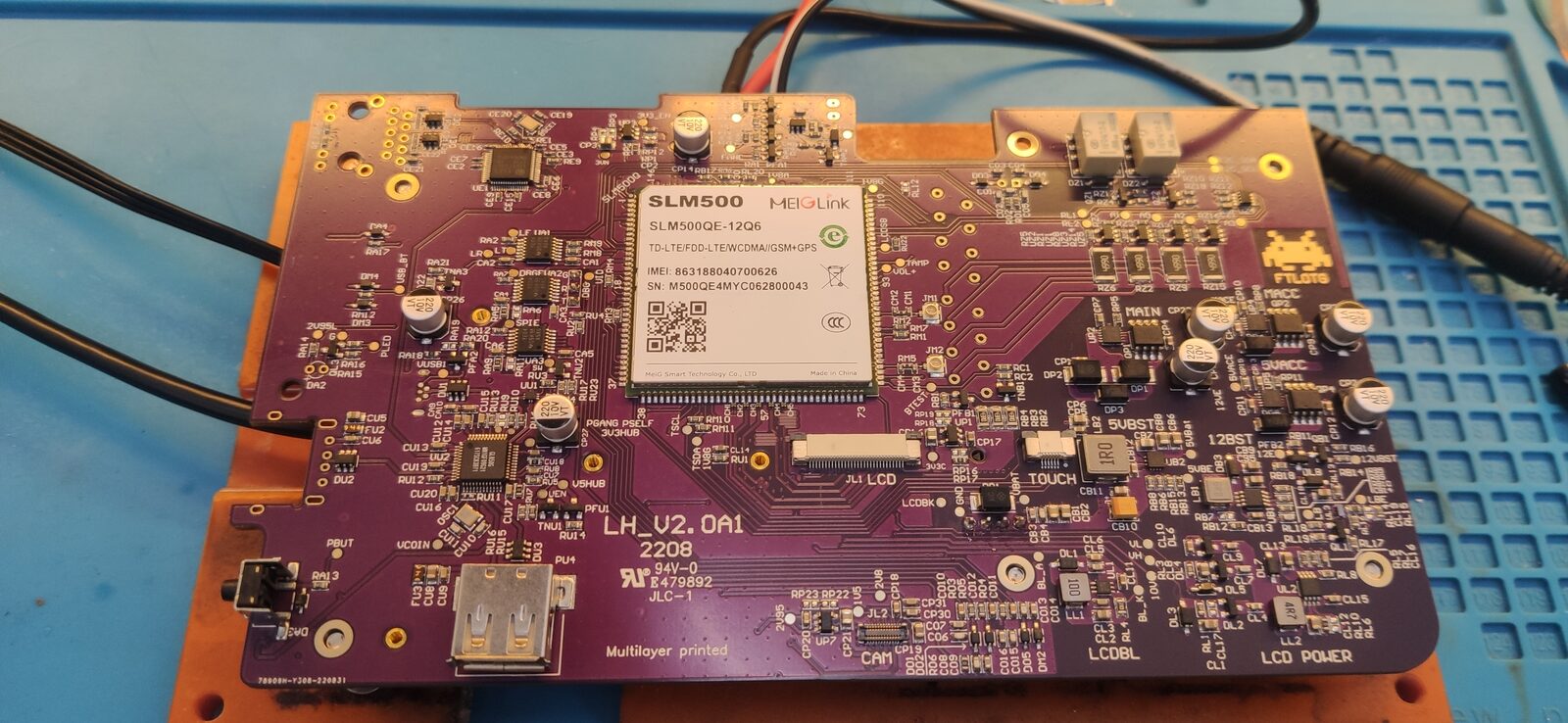 Purple circuit board with a cellular module at its centre and connectors, sitting on a blue work mat with power leads attached.