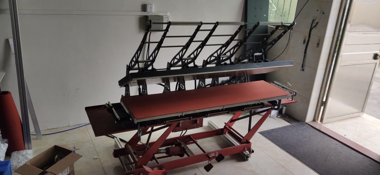 A heat press with a raised upper platen and a red lower bed in a workshop.