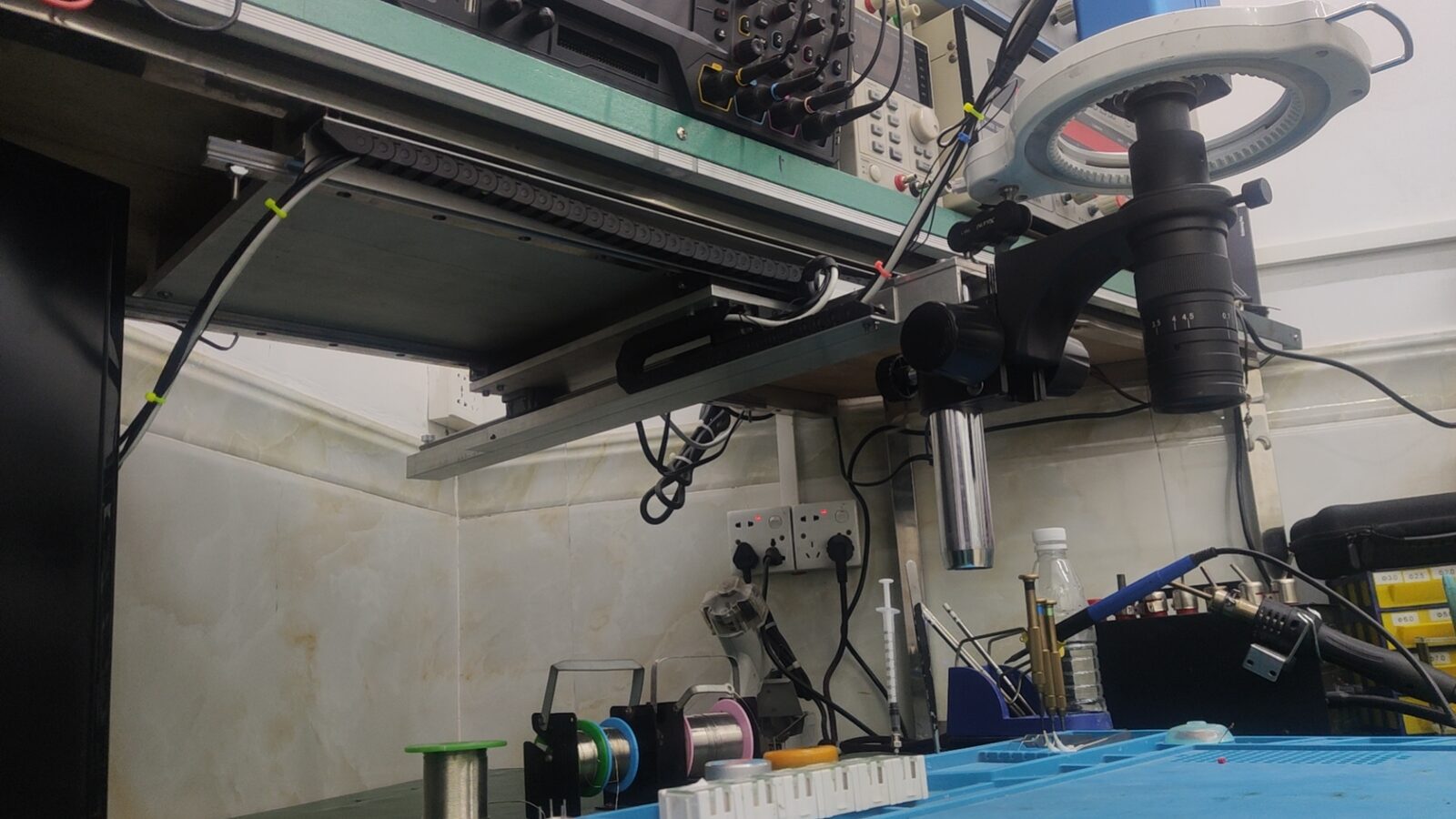 Wide view from the bench level of the microscope with its ring light on the sliding rail, above spools of solder and a blue mat.